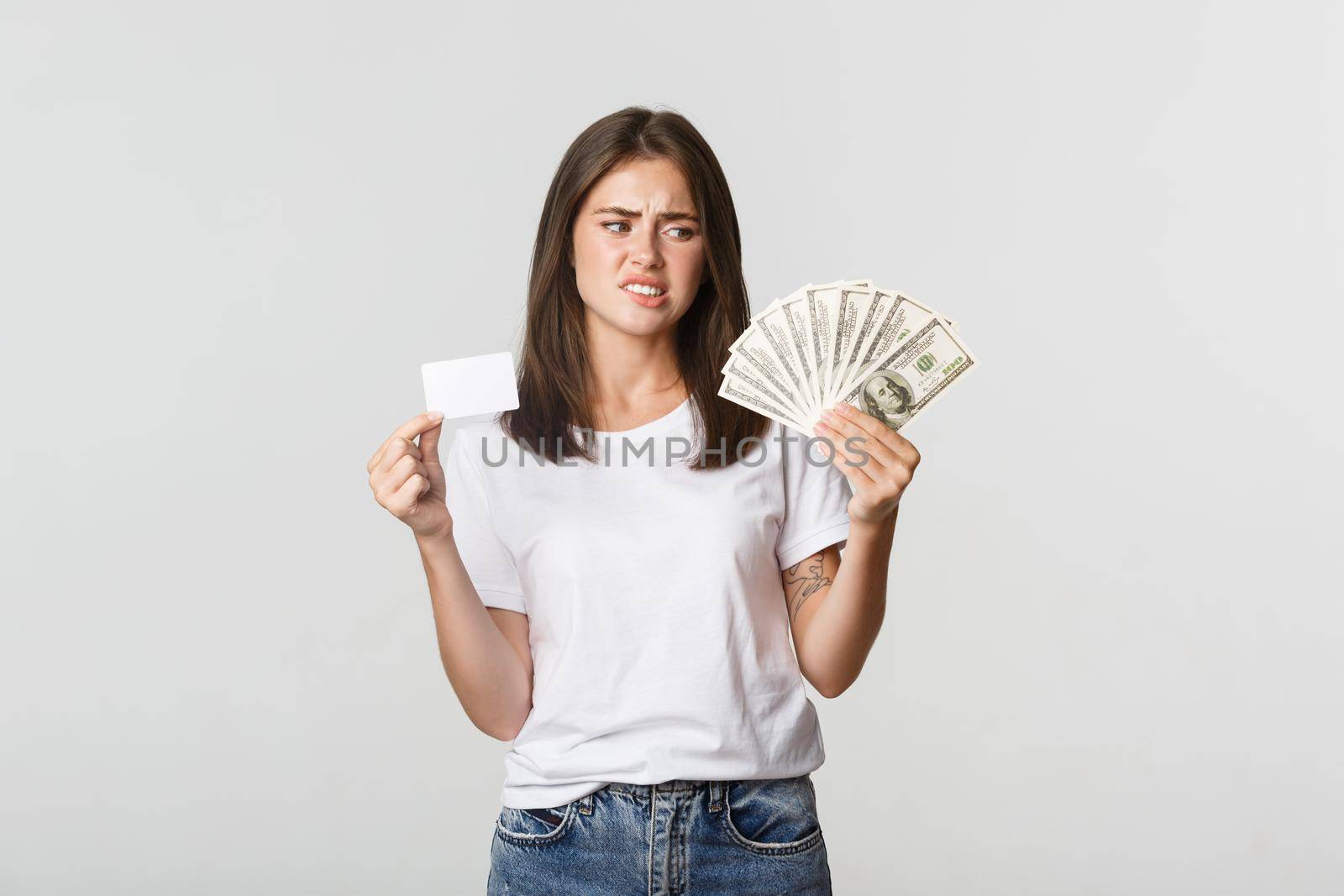 Confused and displeased young woman looking at money and frowning, prefer credit card by Benzoix