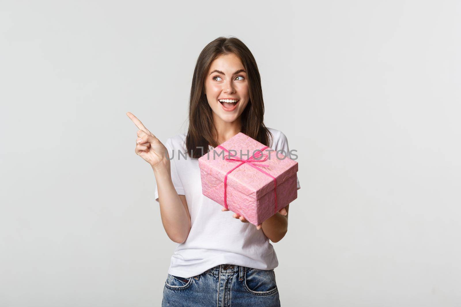Happy smiling brunette girl holding birthday gift and pointing finger left at logo.