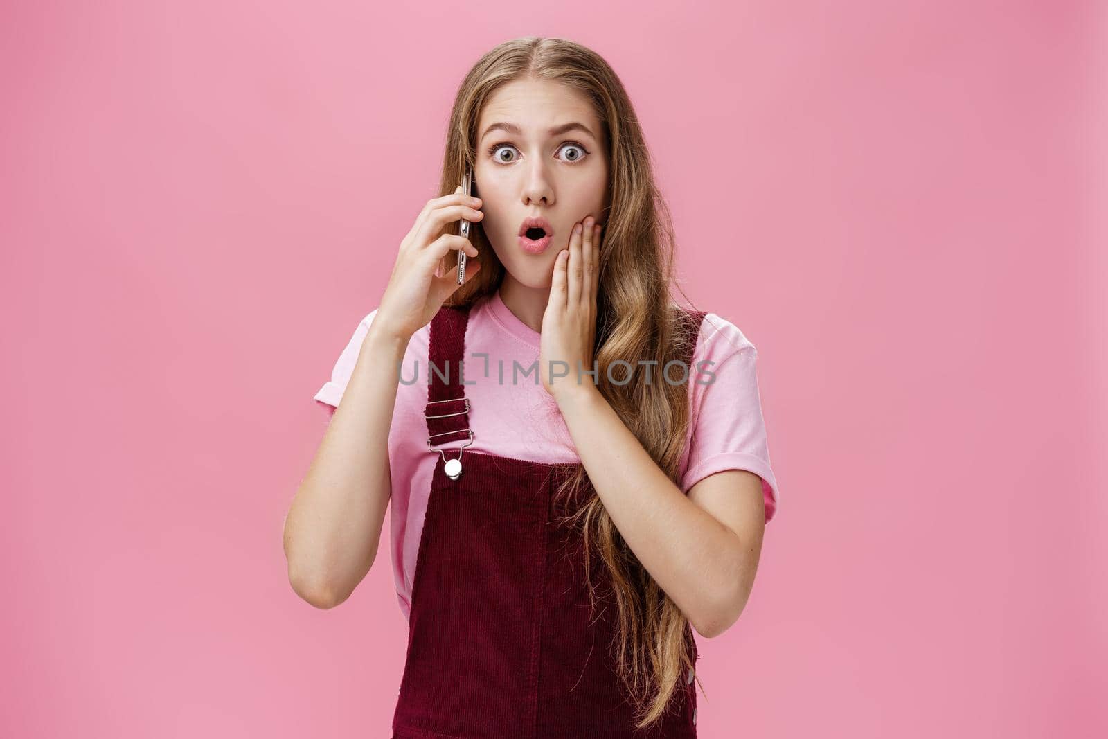 Waist-up shot of shocked woman learning stunning news via smartphone talking on phone holding cellphone near ears folding lips and gasping in surprise popping eyes at camera worried over pink wall.