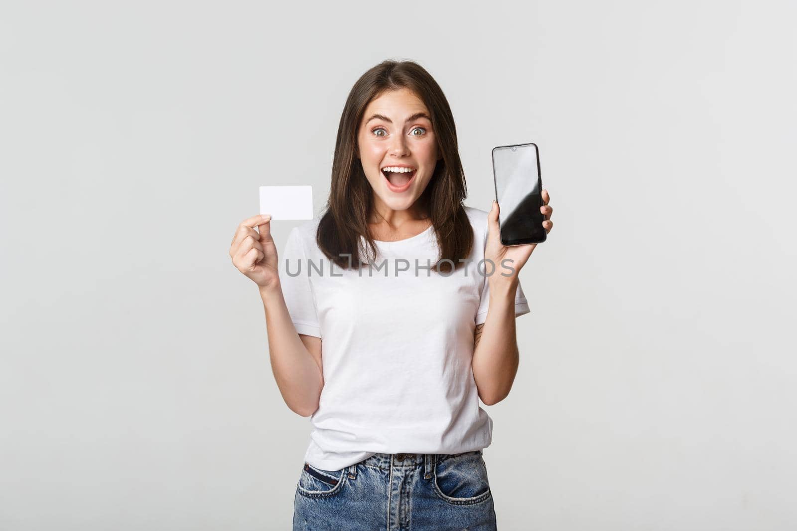 Excited and surprised cute girl showing credit card and mobile phone banking app on screen by Benzoix