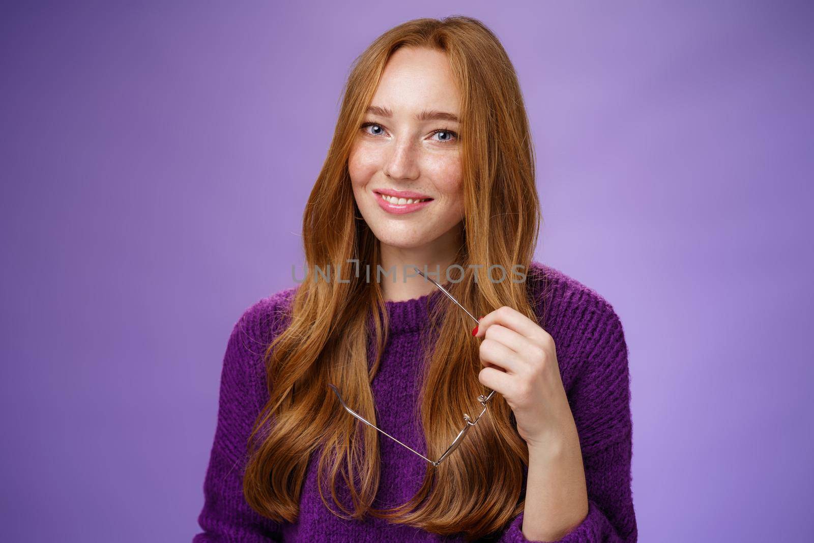Close-up shot of charming tender and cute redhead woman in purple sweater holding glasses and smiling broadly as having nice friendly conversation against violet background. People, facial expression and body language concept