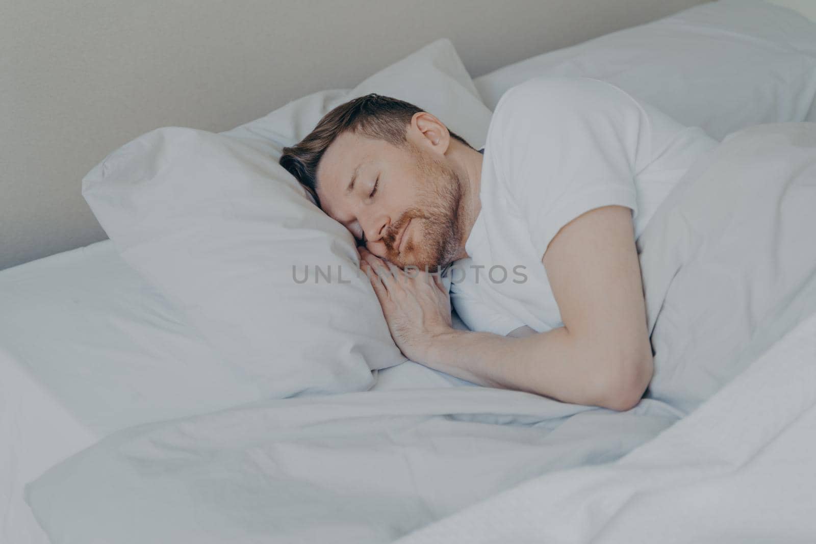Handsome young man in casual domestic clothes with beard comfortably lying and sleeping in cozy bed with white beddings at home, male resting after hard working day. Bedtime concept