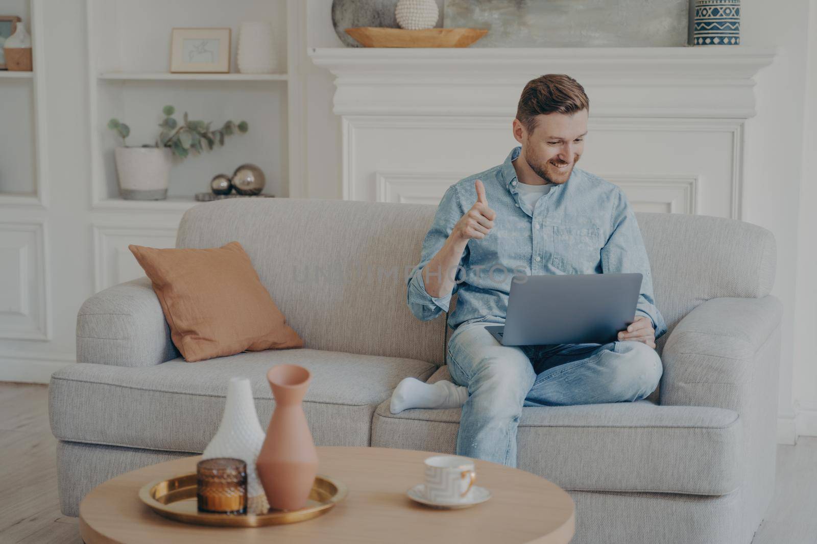 Young male business worker sitting on couch with crossed leg, showing thumbs up gesture to his co-workers in online meeting, displying his satisfaction with completed work