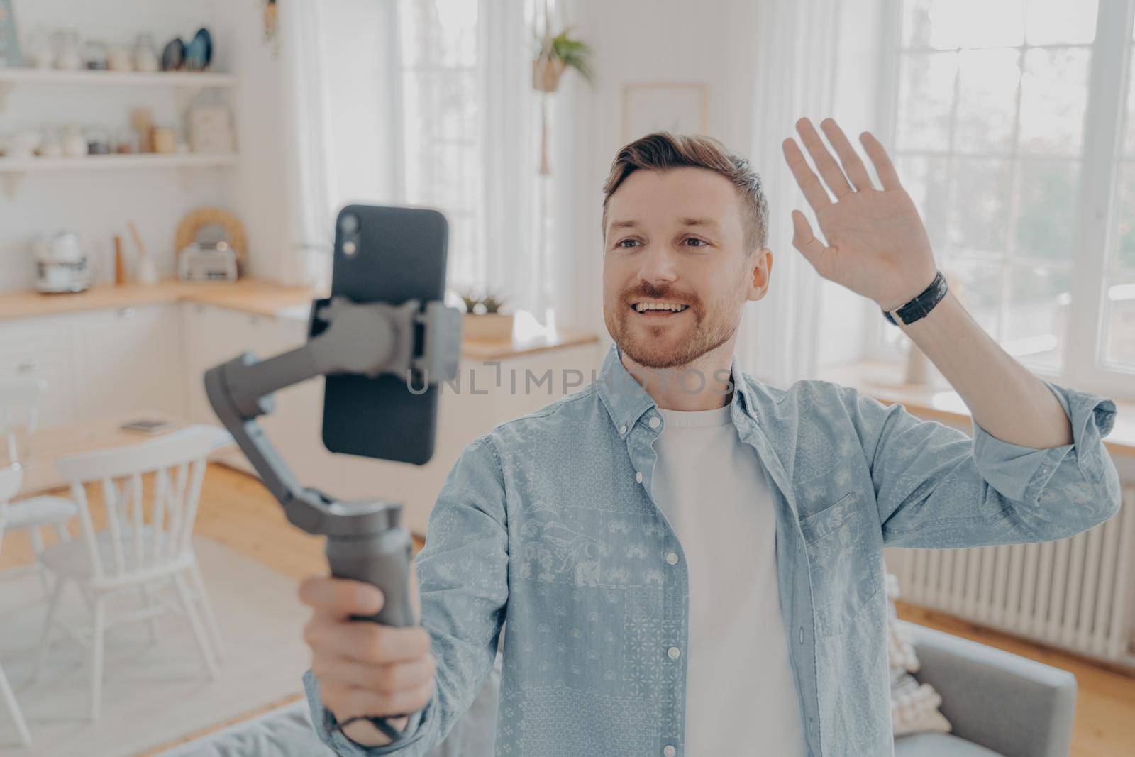 Domestic handsome guy blogger using smartphone with gimbal, waving hand in hello gesture and telling followers in social media about his life while standing in living room at home