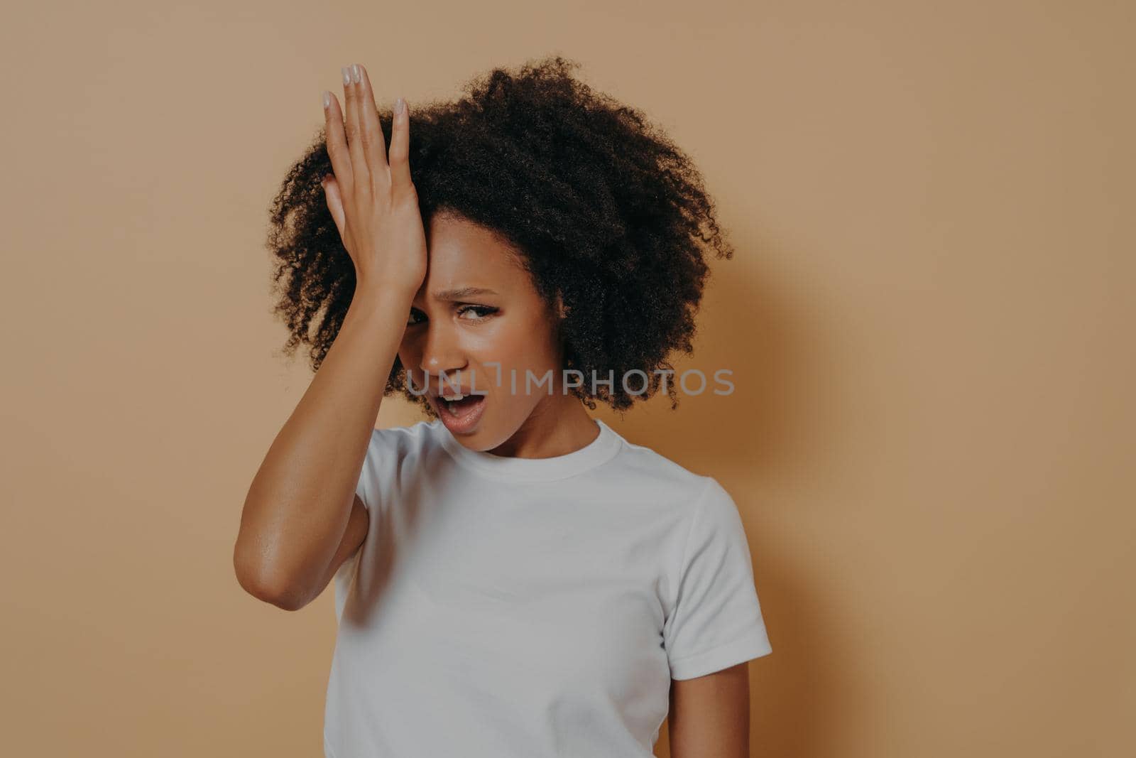 Studio shot of young stressful african woman with hand on forehead suffering from strong headache or head migraine, looking at camera with unpleased face expresion, isolated on sand color background