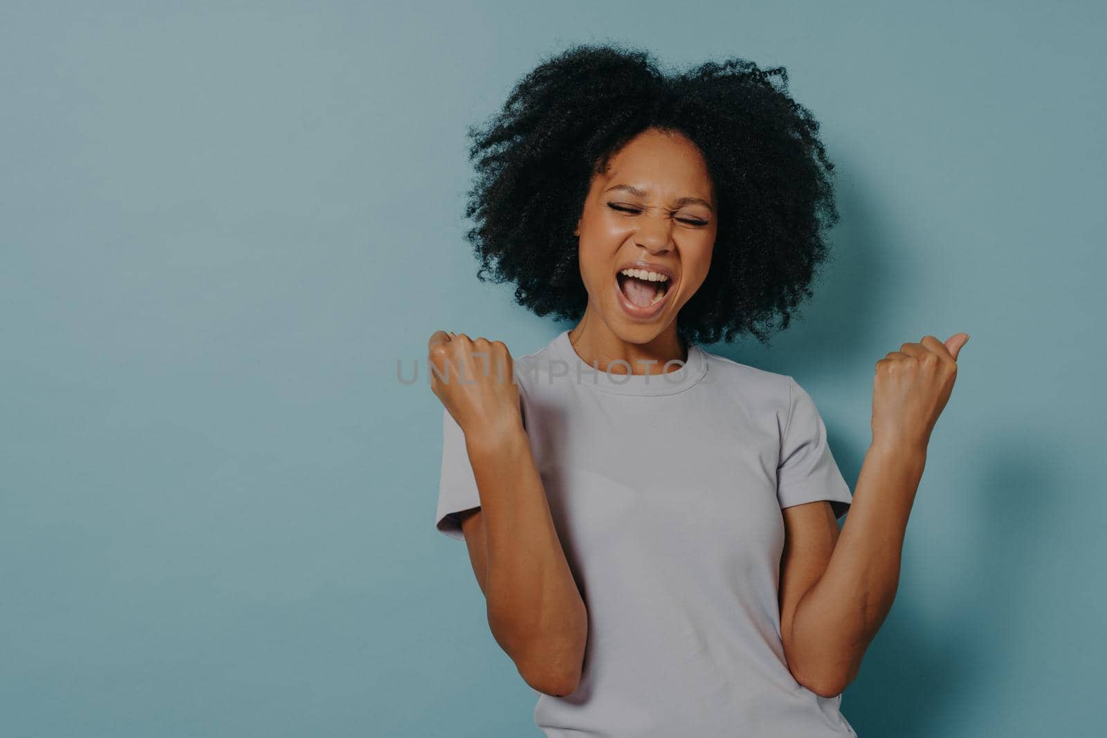 Overjoyed girl clenches fists with happiness, widely opens mouth as shouts loudly. Photo of african american girl wears casual outfit on blue studio background. Emotions and positive feelings concept