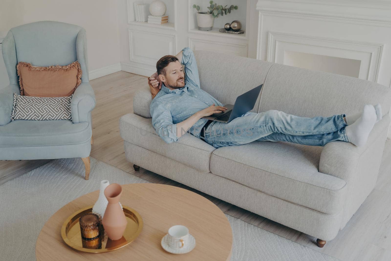 Young businessman working remotely at home while lying on couch by vkstock