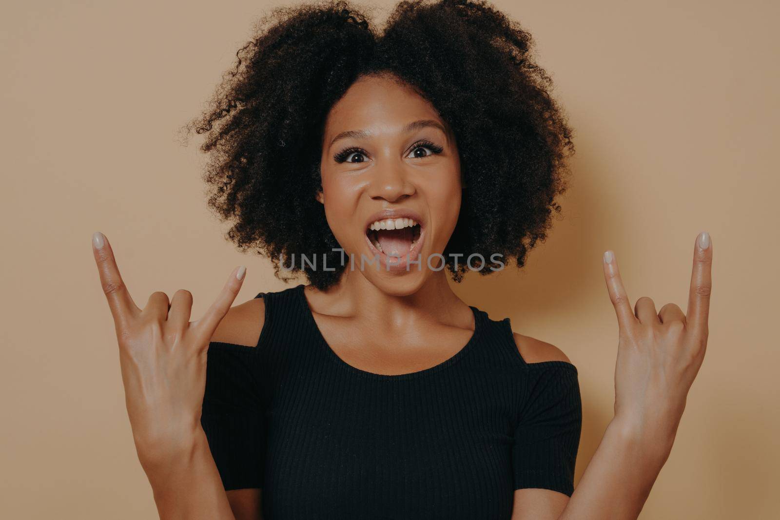 Young african american woman wearing black tshirt shouting with crazy facial expression doing rock-n-roll symbol with hands up like music star, isolated over beige studio background with copy space