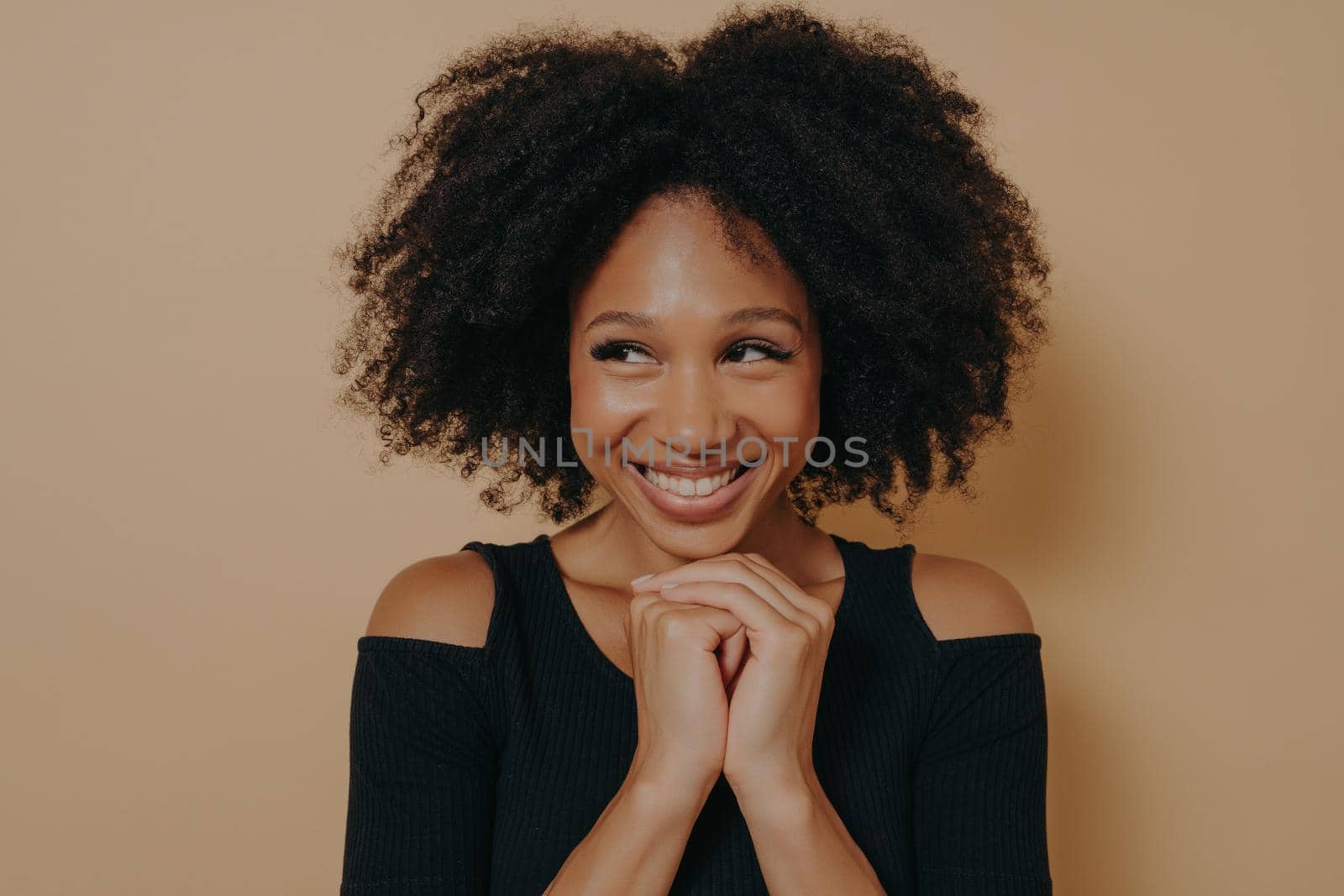 Studio shot of happy Afro girl keeps hands together under chin, being in high spirit, likes friends idea for next day, isolated on dark beige wall background with copy space. Positive emotions concept