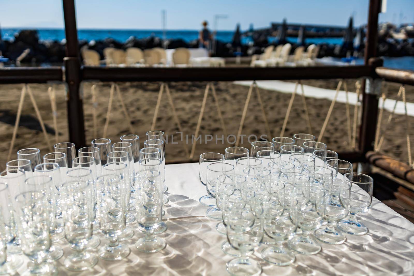 Table full of champagne and wine glasses in front of preparations for wedding party on a beach by Wierzchu