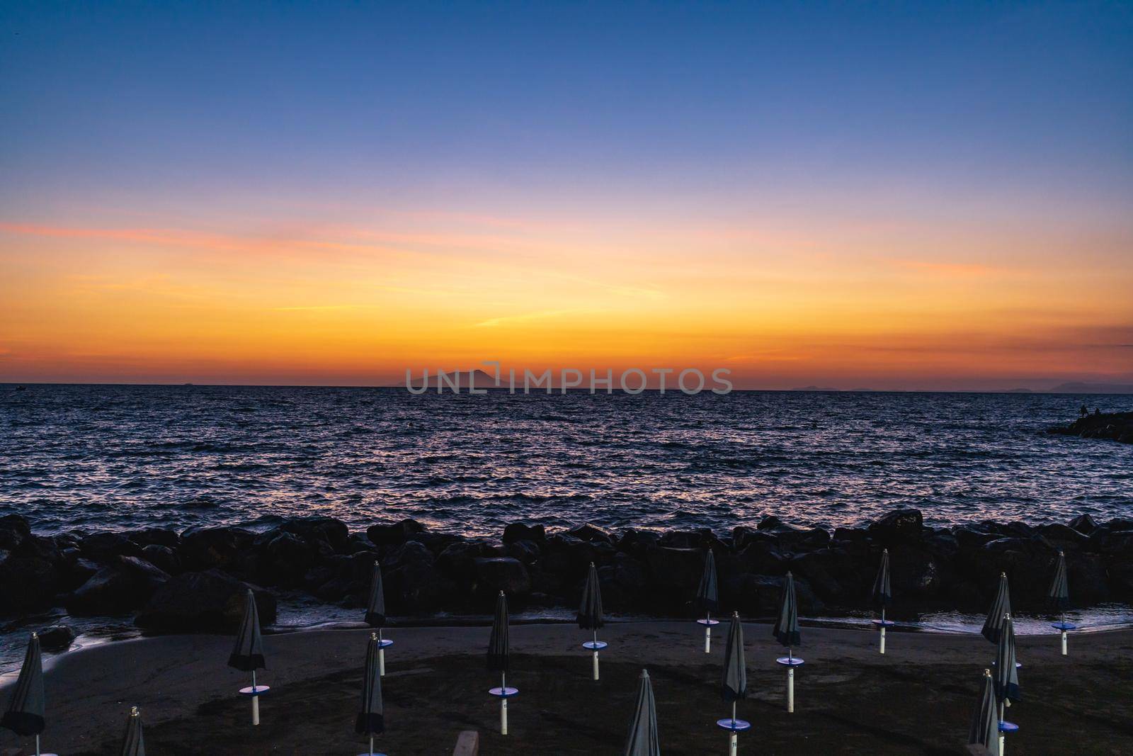 Beautiful sunset over the sea and small beach full of blue and white umbrellas next to giant rocks