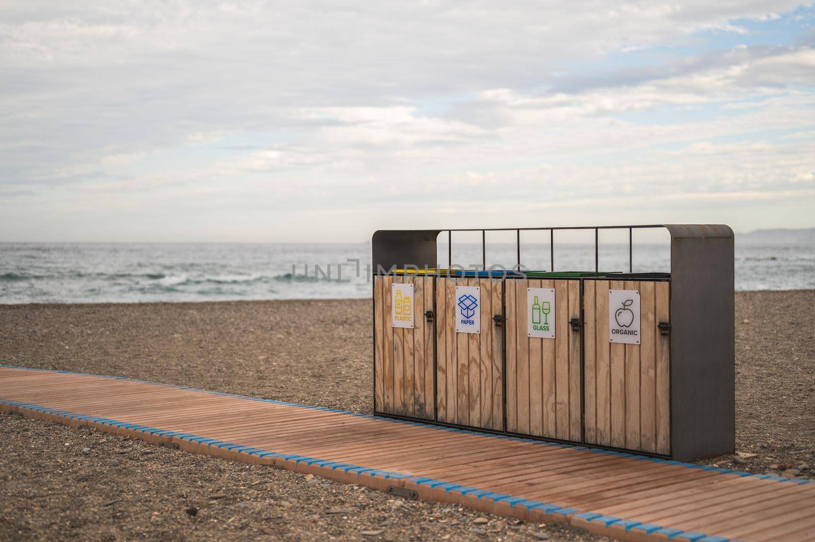 recycling containers in front of a wooden path on the beach with the sea behind and cloudy sky. concept of conservation of the environment