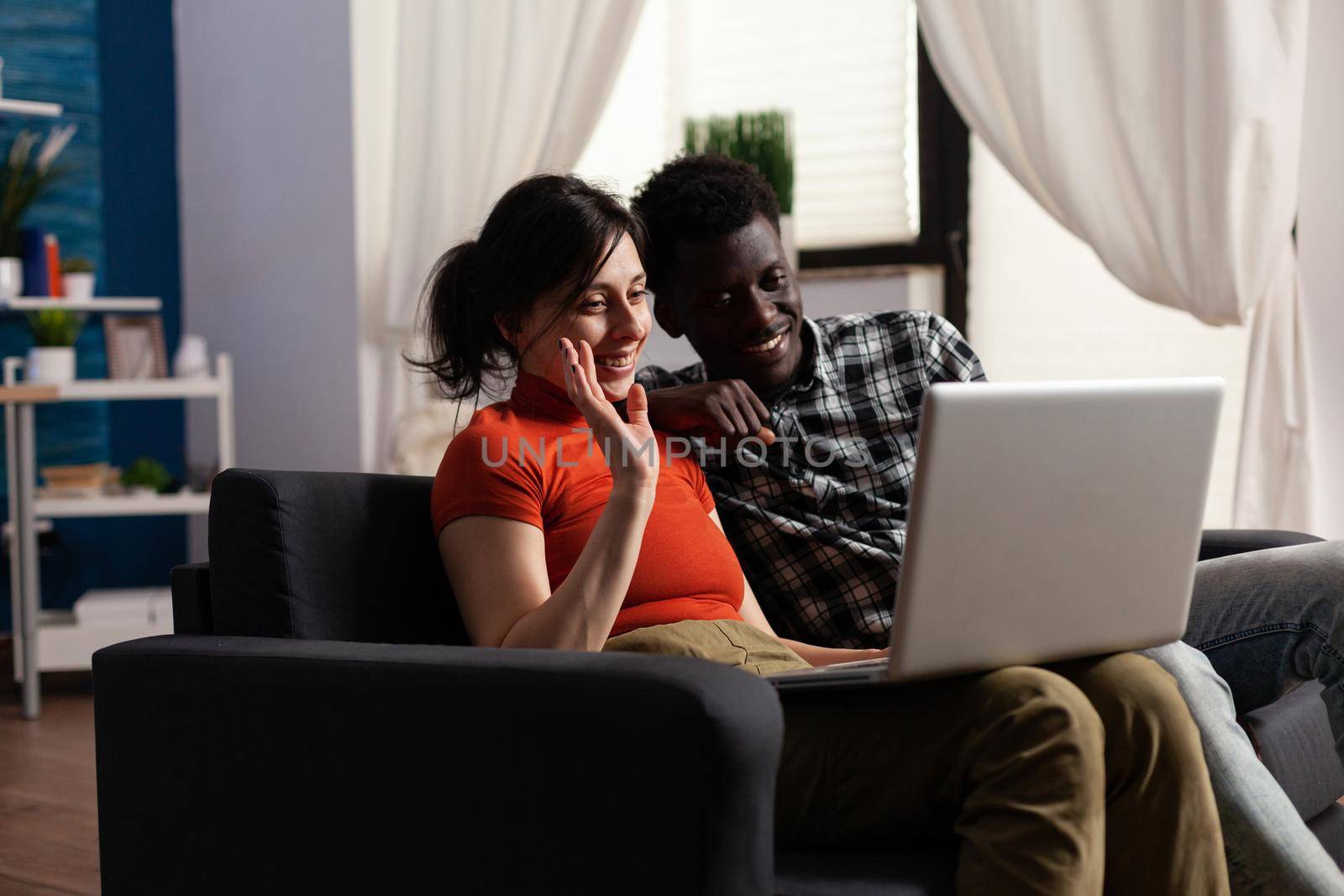 Interracial couple using video call communication on laptop at home. Mixed race people waving at camera while talking to friends on online remote conference via internet connection