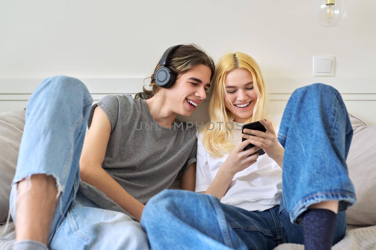 Couple of cheerful emotional teenagers friends having fun together, sitting on couch looking into smartphone. Adolescents male and female 15, 16 years old, technology, lifestyle, fun, friendship