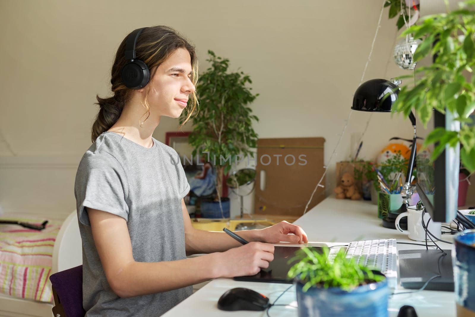 A teenage guy in headphones sitting at a table at home using a computer and a graphics tablet. Creativity, hobby, leisure, adolescence, technology, school, learning concept