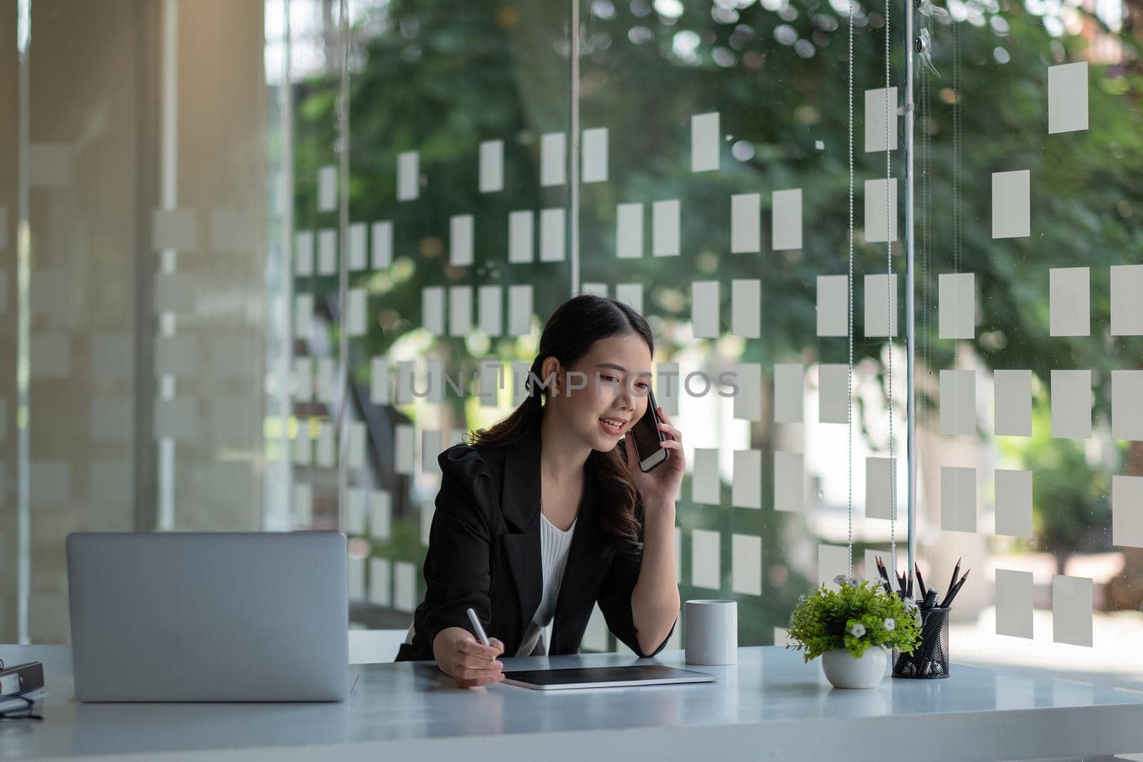 Smiling young asian businesswoman sit at desk smiles talking on smartphone looking out the window enjoy pleasant conversation with client, female sales manager makes successful profitable deal concept.