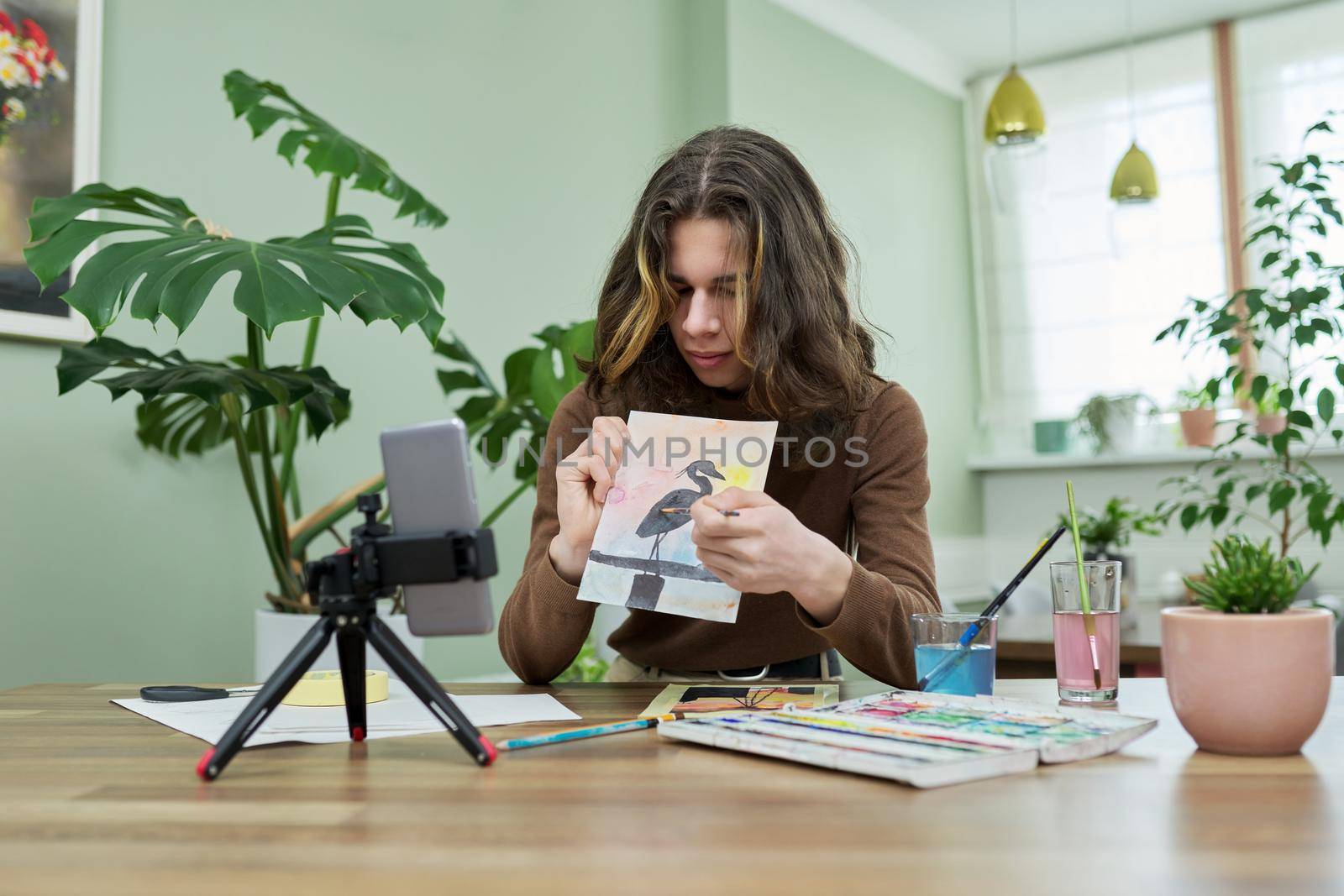 Guy teenager student drawing online using smartphone. Teenage male with watercolors, brushes, sketch, sitting at home, showing picture on phone. Video lesson, virtual meeting with teacher, creativity