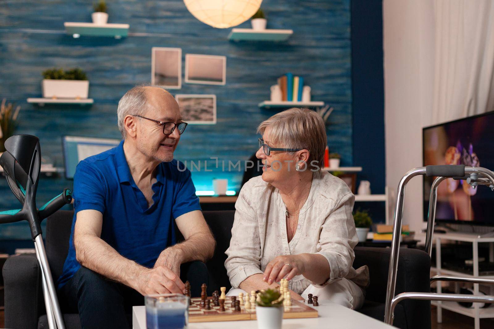 Senior couple playing chessboard game on table in living room. Retired husband and wife enjoying fun activity together on sofa. Married elderly people with wheelchair, crutches and walk frame