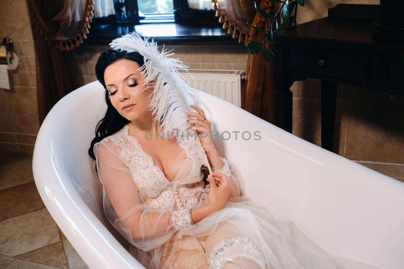 the bride, dressed in a boudoir transparent dress and underwear, lies in a vintage bathroom with a white feather in her hands, The morning of the bride.