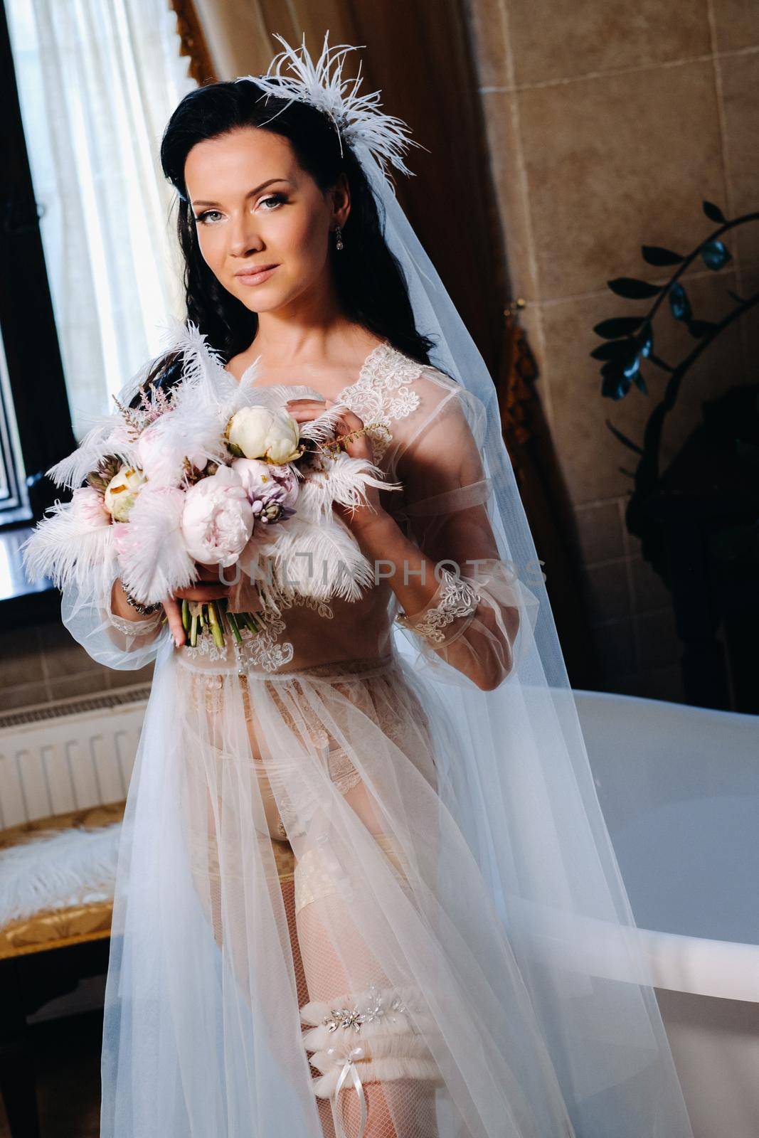 the bride, dressed in a boudoir transparent dress and underwear, sits near a vintage bath with a feather in her hands, The morning of the bride.