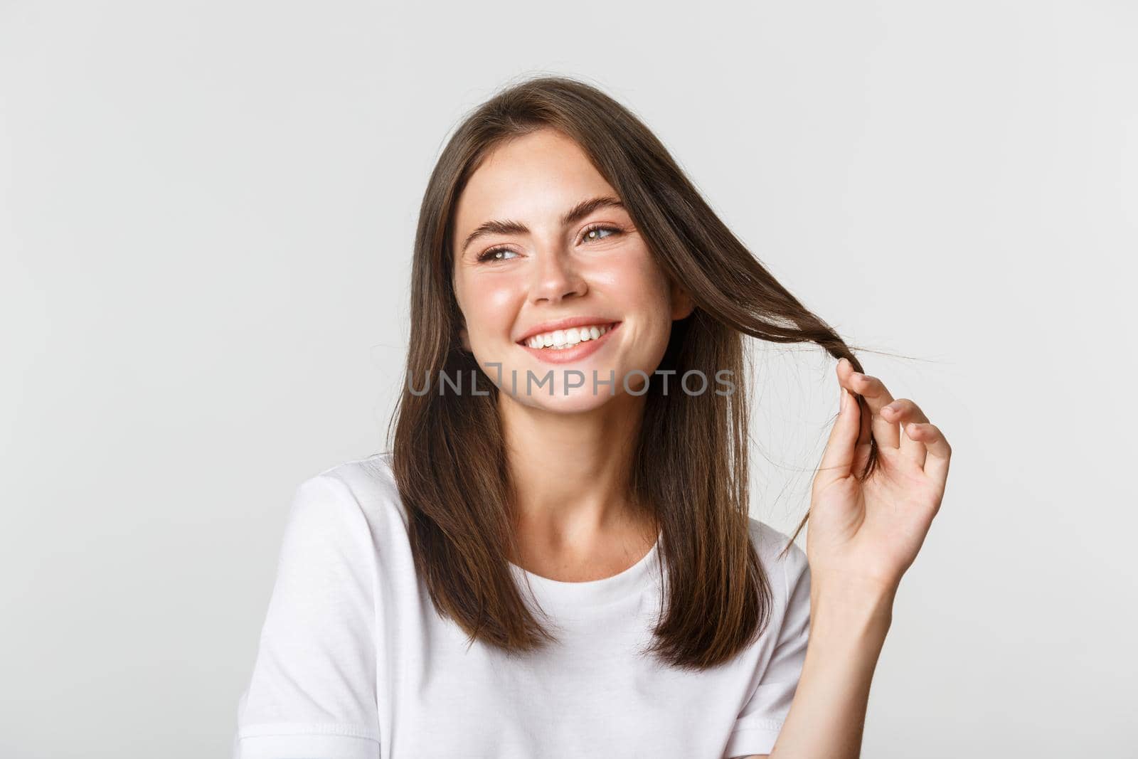 Portrait of flirty beautiful girl playing with hair and smiling at camera coquettish, standing white background.