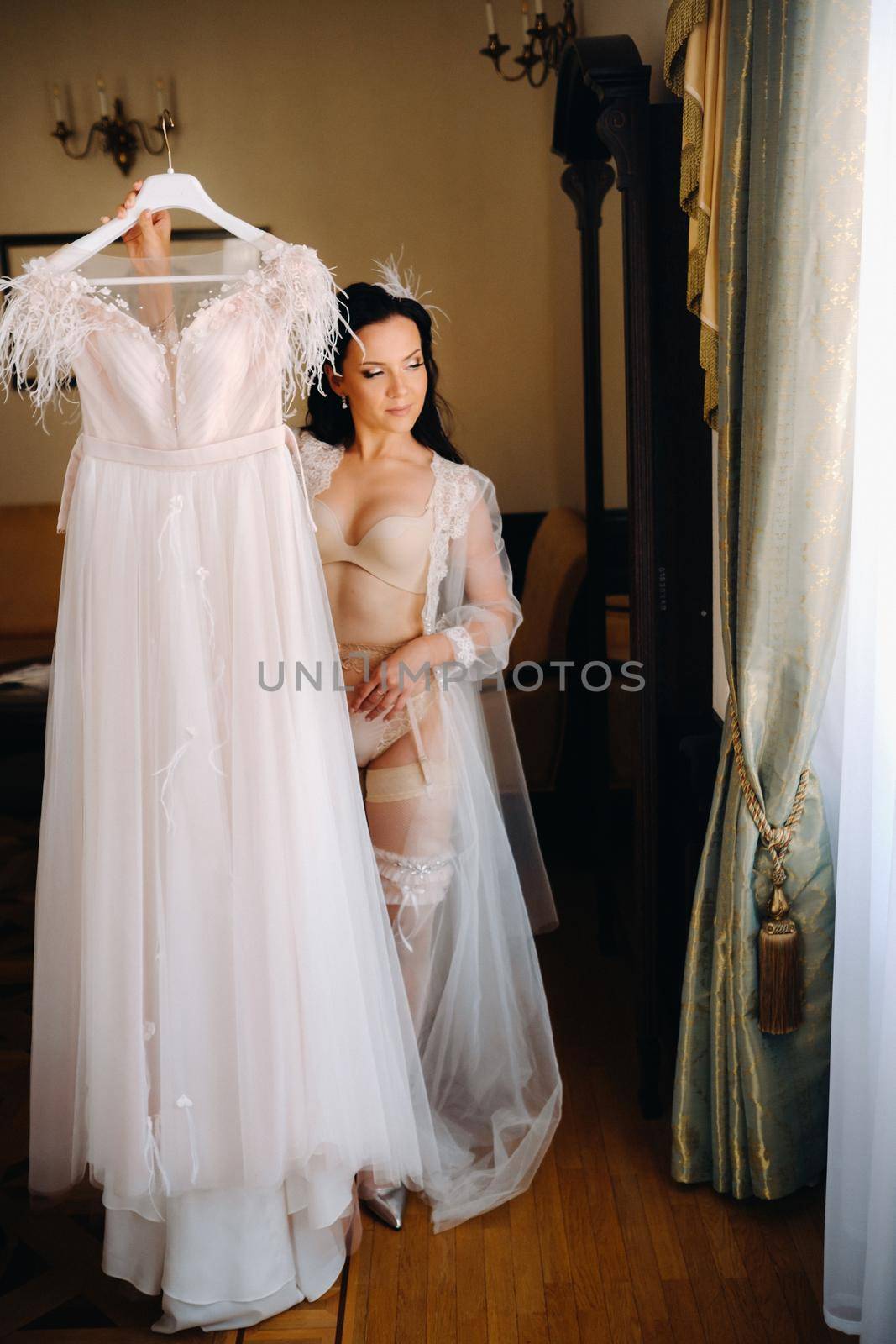 a bride dressed in a boudoir transparent dress and underwear holds her wedding dress in her hands in the interior of the house.