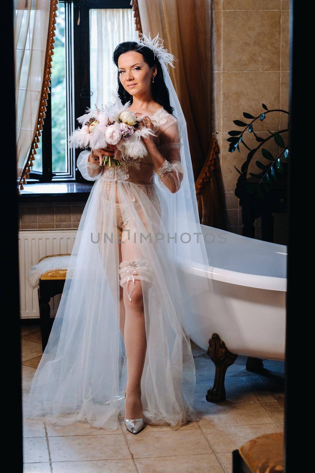 the bride, dressed in a boudoir transparent dress and underwear, sits near a vintage bath with a feather in her hands, The morning of the bride.