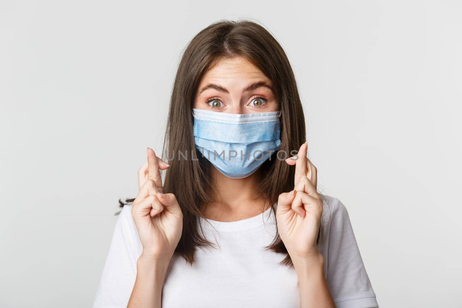 Covid-19, health and social distancing concept. Close-up of hopeful smiling girl in medical mask, cross fingers good luck.