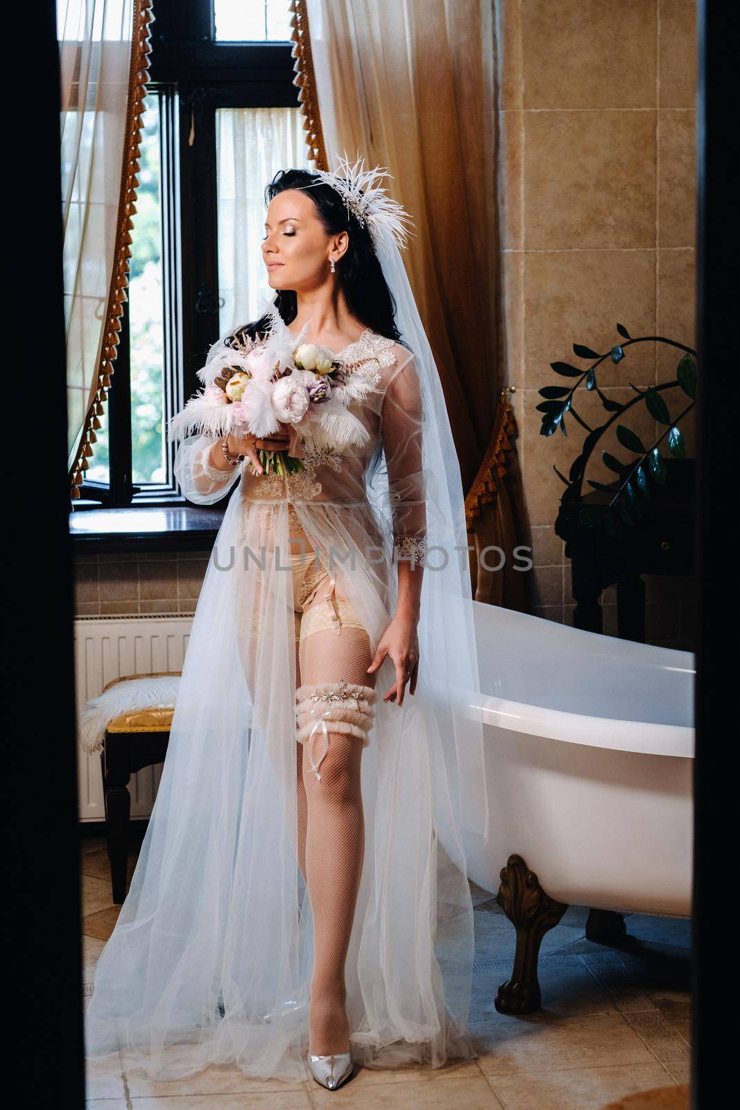 the bride, dressed in a boudoir transparent dress and underwear, sits near a vintage bath with a feather in her hands, The morning of the bride.