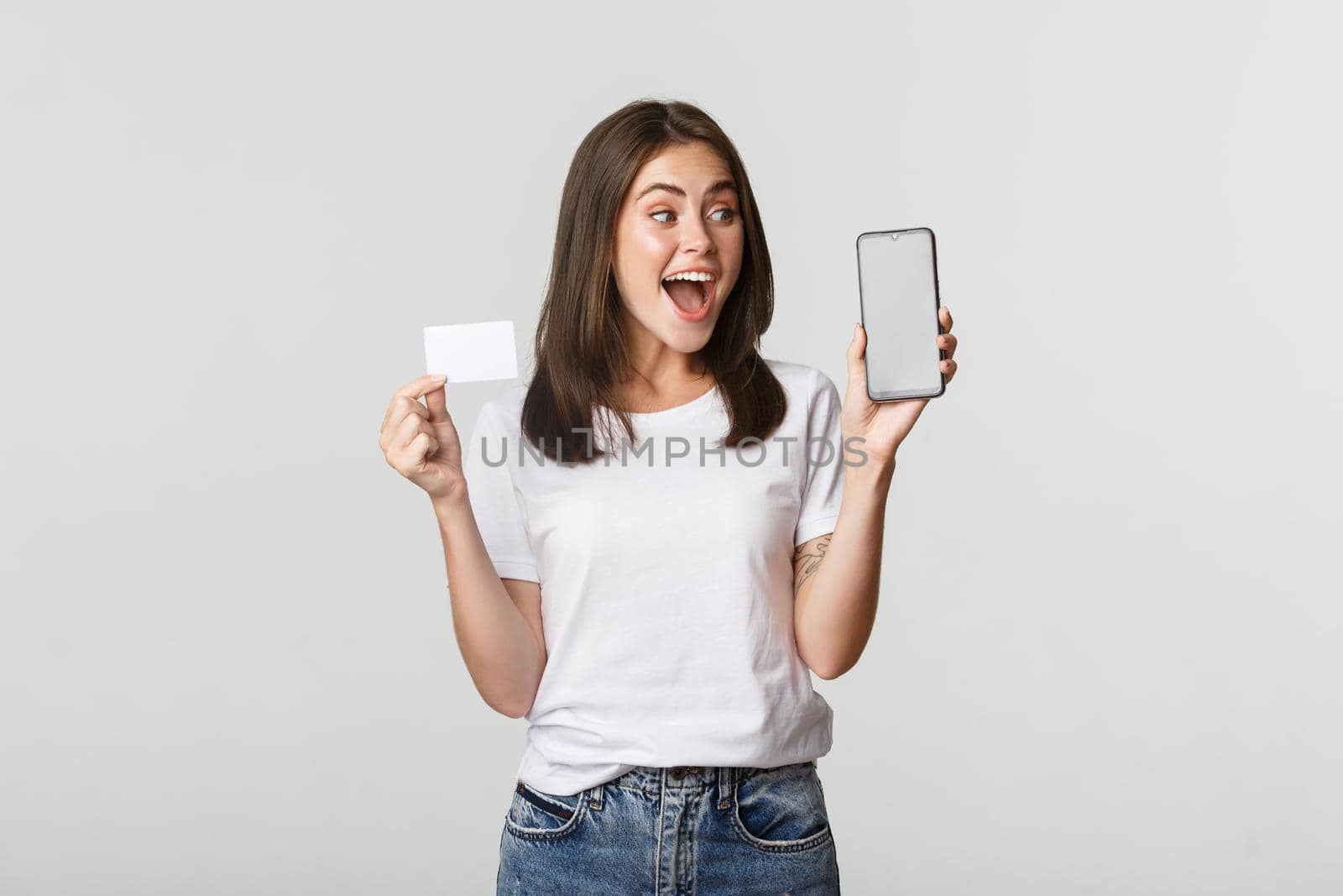 Excited and surprised cute girl showing credit card and mobile phone banking app on screen.
