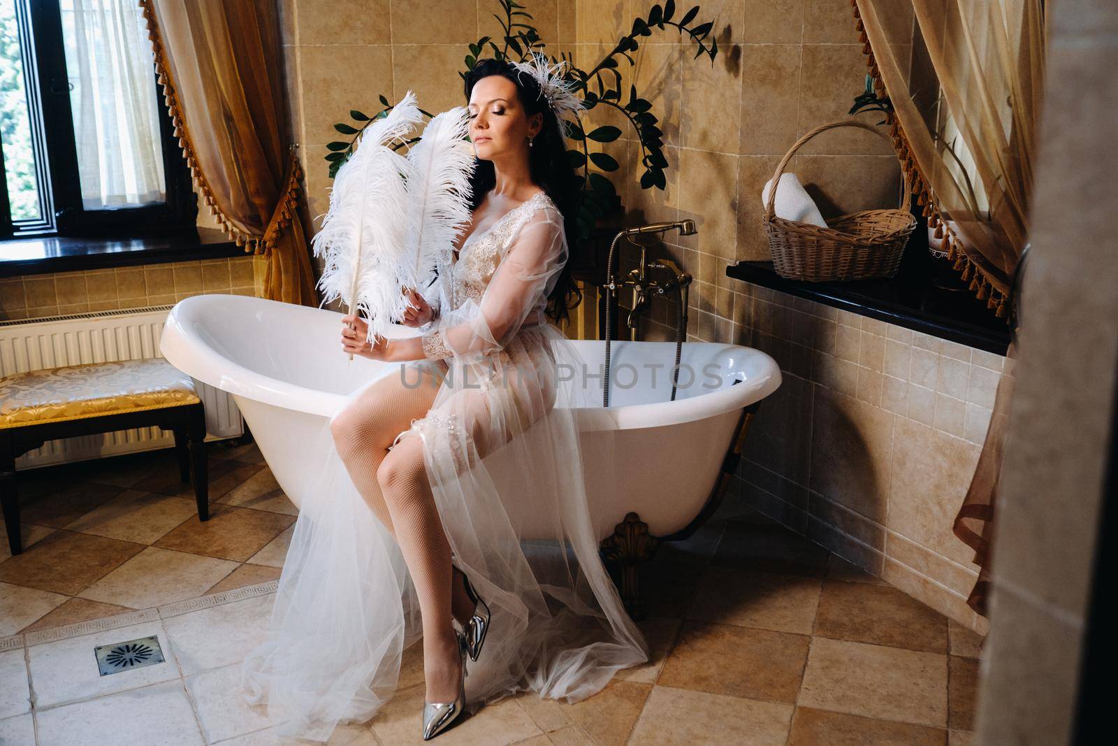 the bride, dressed in a boudoir transparent dress and underwear, sits on a vintage bathtub with a feather in her hands, The morning of the bride.