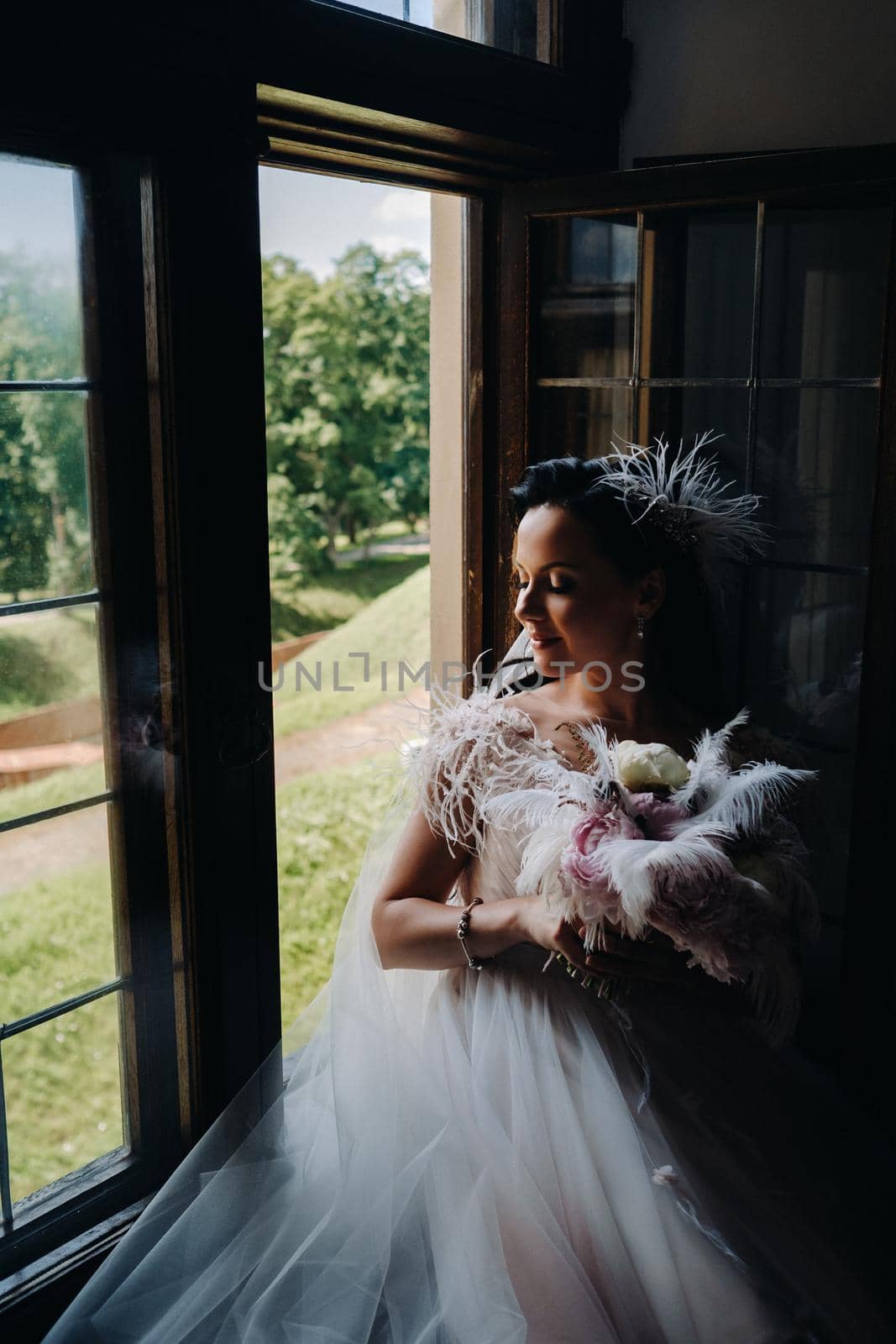 A bride in a wedding dress and a bouquet sits at an open old window and looks.