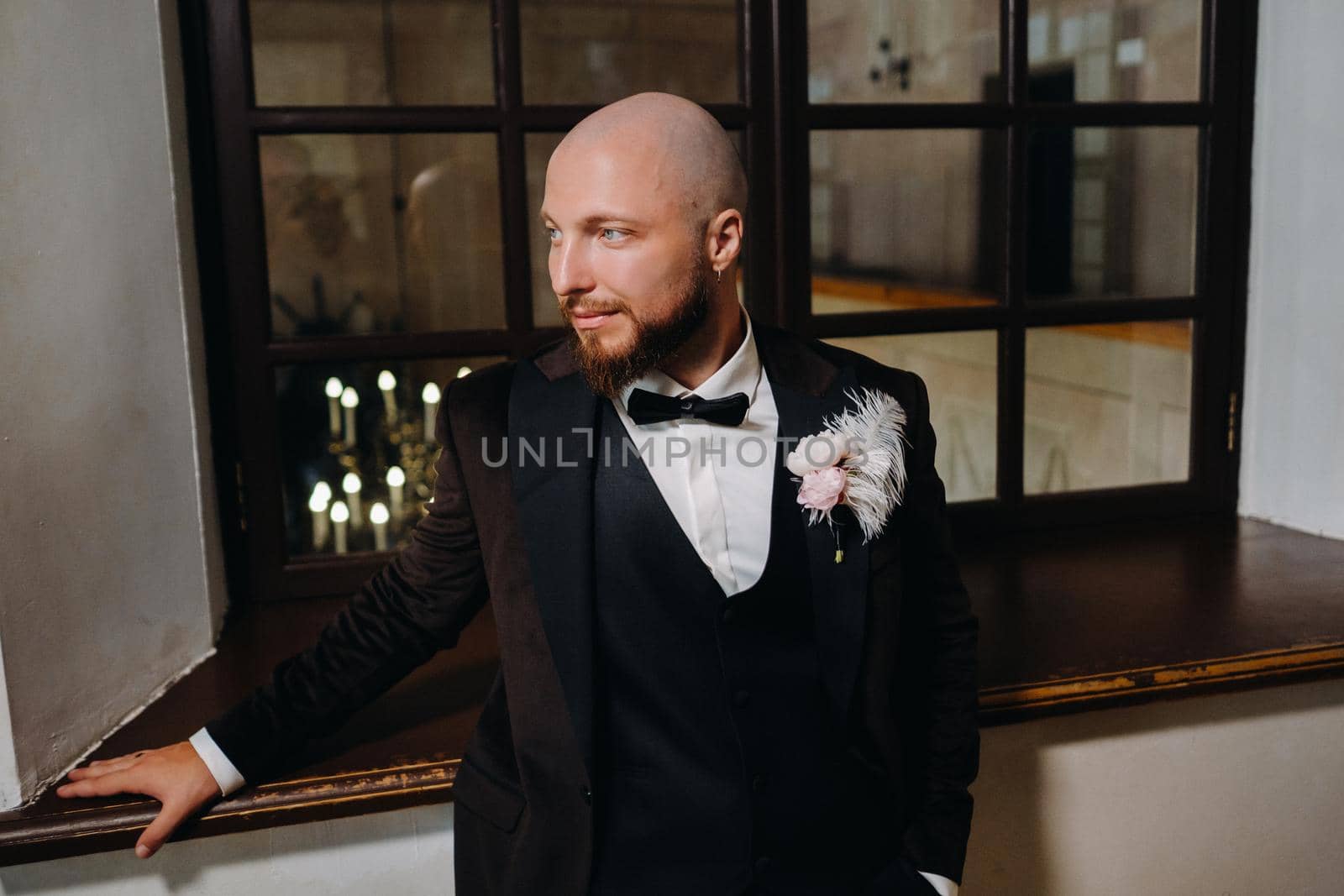 Portrait of the groom in a suit and bow tie, standing in the interior.