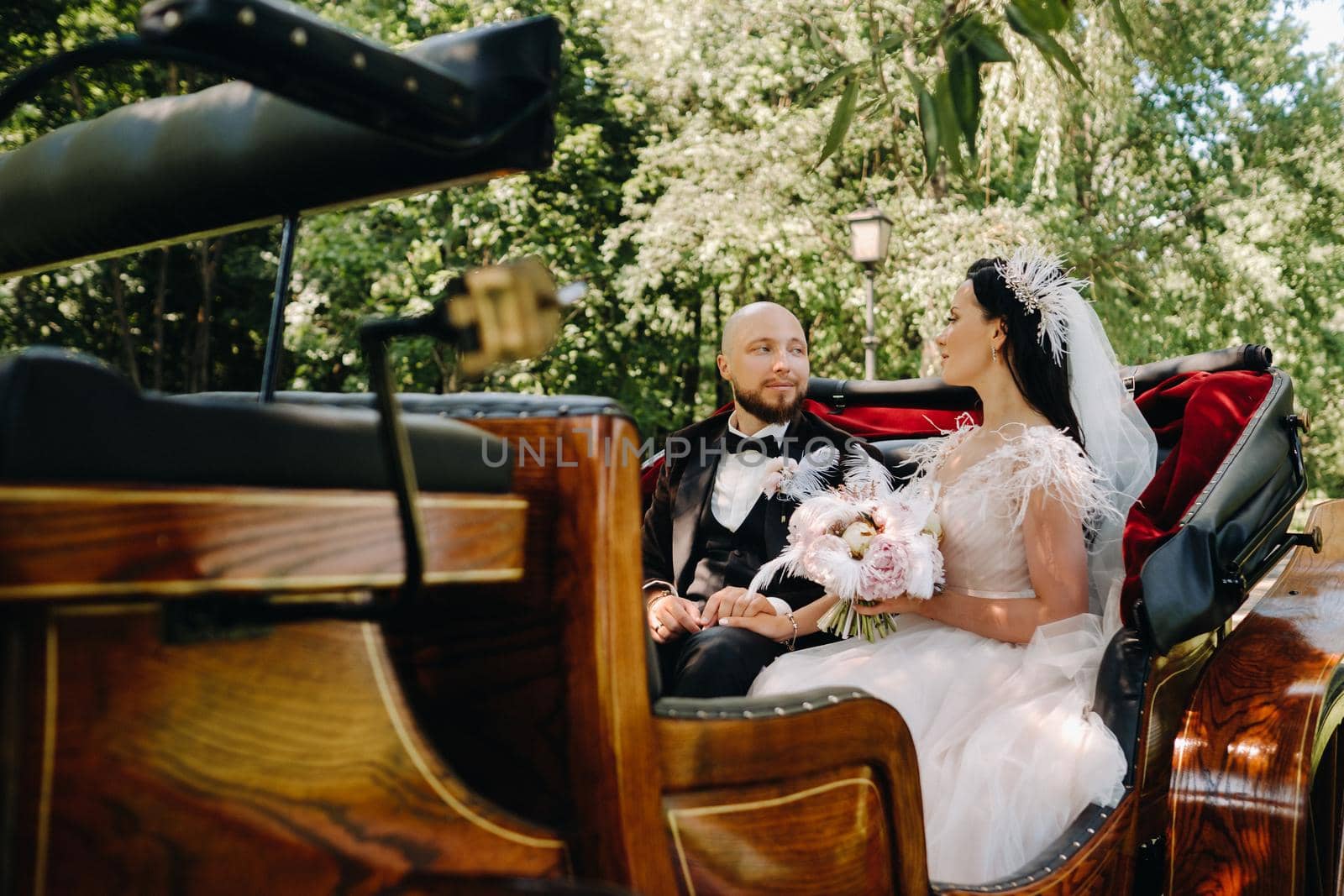 The bride and groom with a bouquet are sitting in a carriage in nature in retro style by Lobachad
