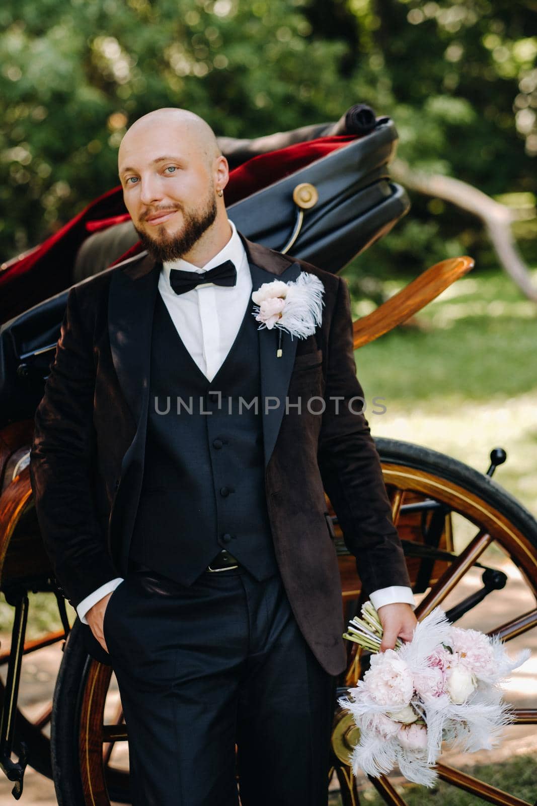 The groom with a bouquet in his hands stands near the carriage in nature in retro style by Lobachad