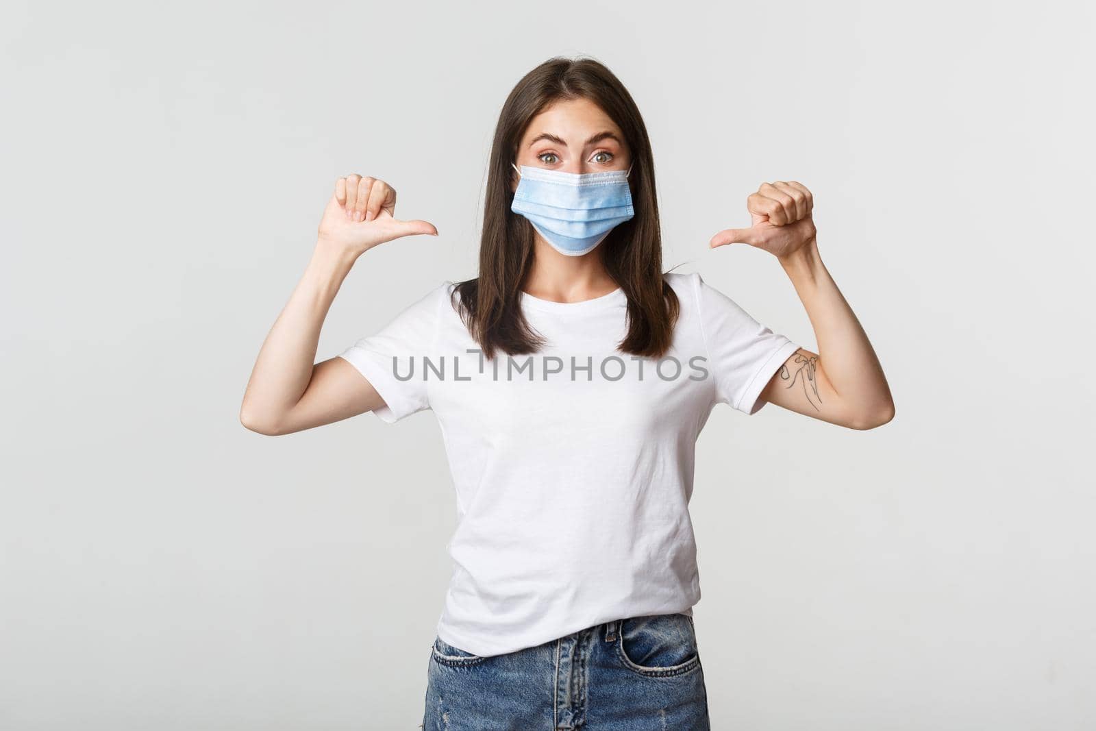 Covid-19, health and social distancing concept. Attractive smiling woman in medical mask pointing at herself, white background.