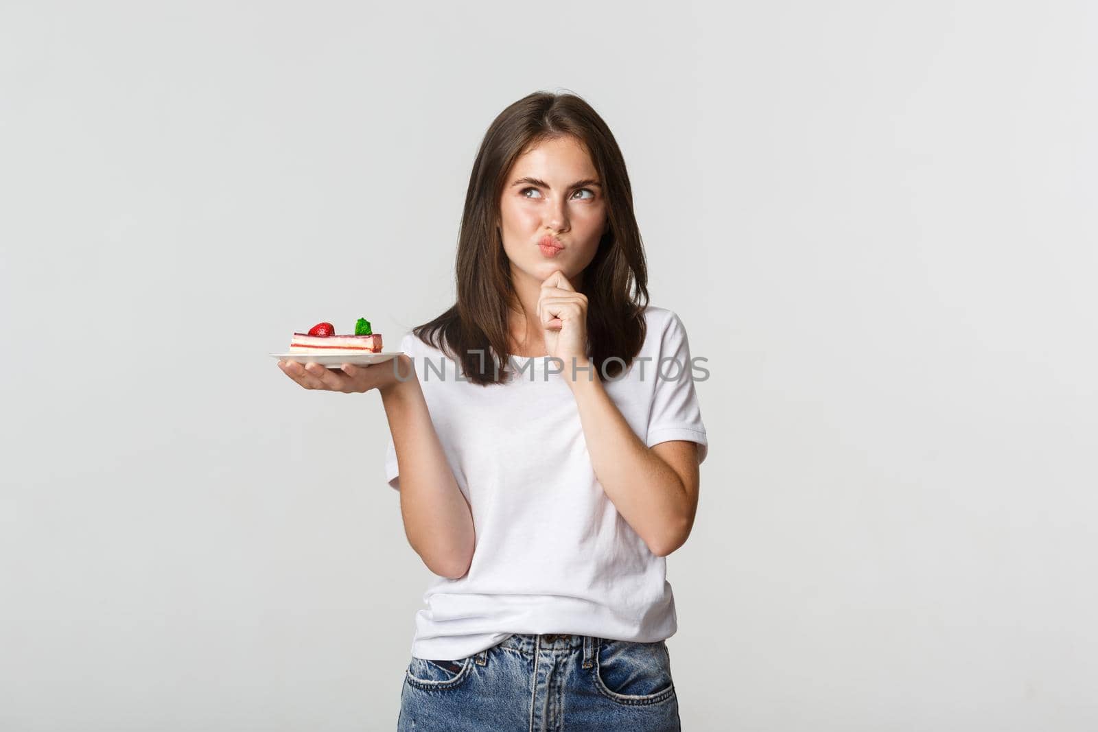 Thoughtful smiling pretty girl pondering while holding piece of cake, white background.