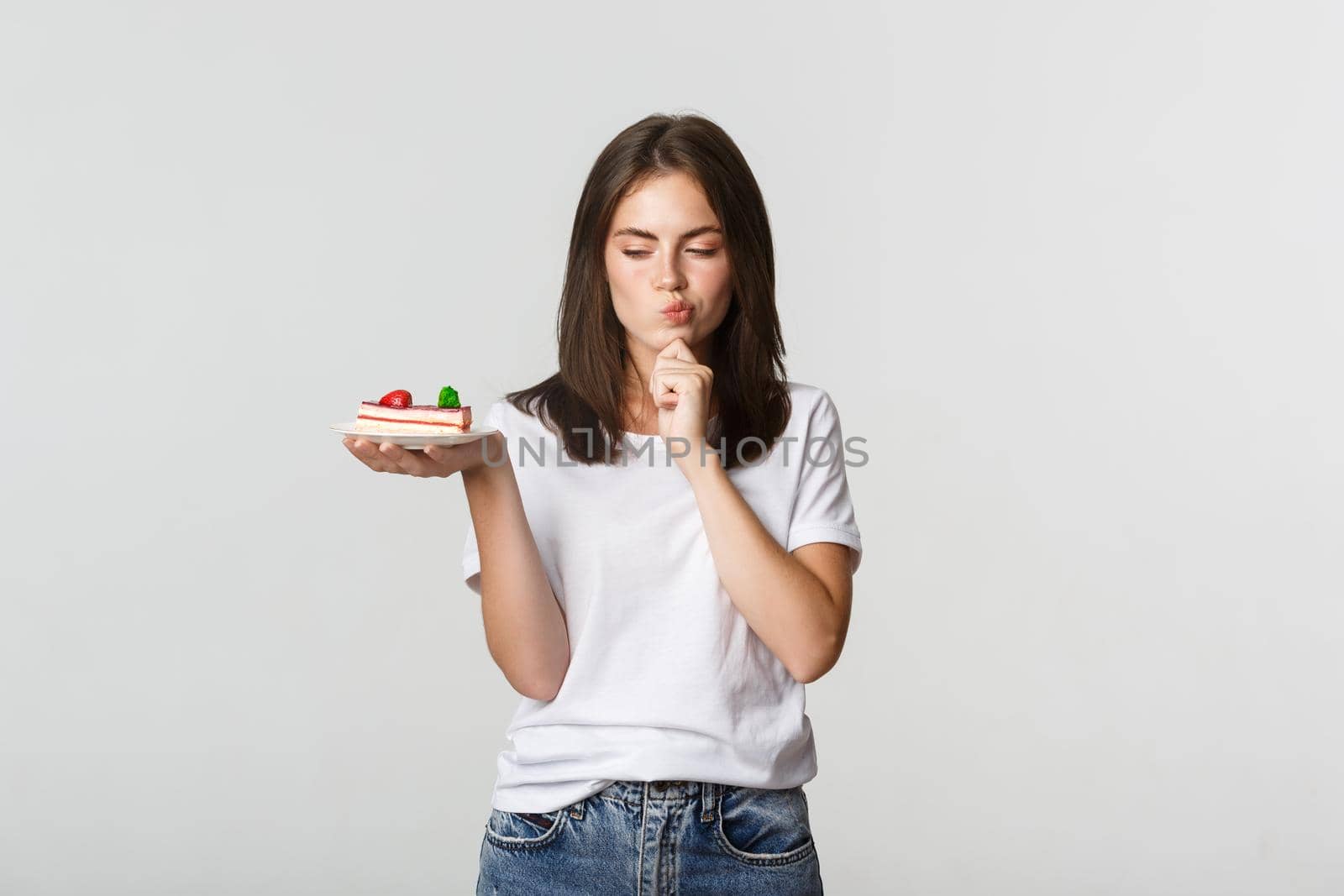Thoughtful smiling pretty girl pondering while holding piece of cake, white background.