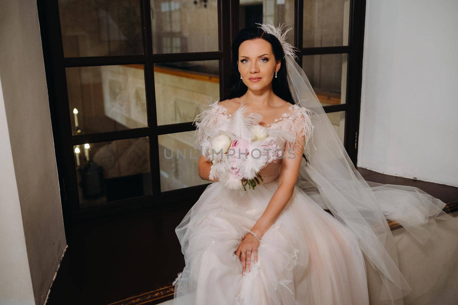 A bride in a wedding dress and a bouquet sits at an open old window and looks.