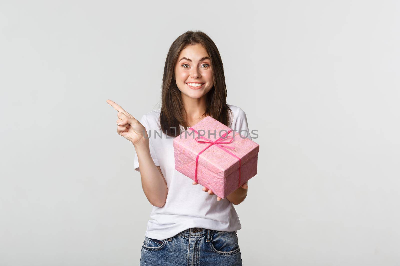 Happy smiling brunette girl holding birthday gift and pointing finger left at logo.