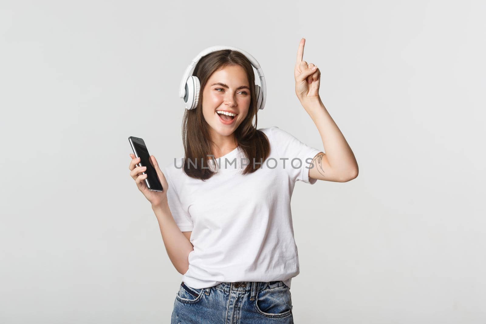 Happy beautiful brunette girl dancing and listening music in wireless headphones, holding smartphone by Benzoix