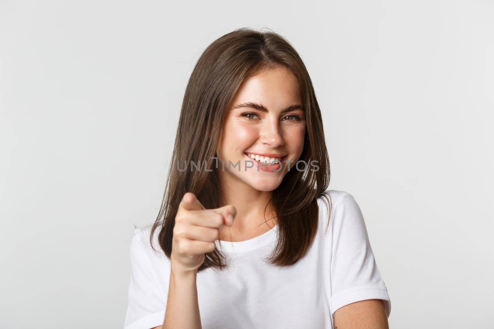 Close-up of happy smiling brunette girl pointing at camera, standing white background.