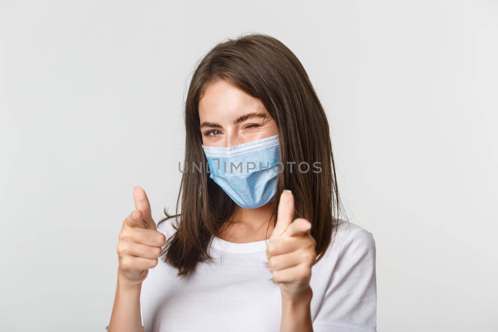 Covid-19, health and social distancing concept. Sassy smiling brunette girl in medical mask winking flirty at camera and pointing fingers.