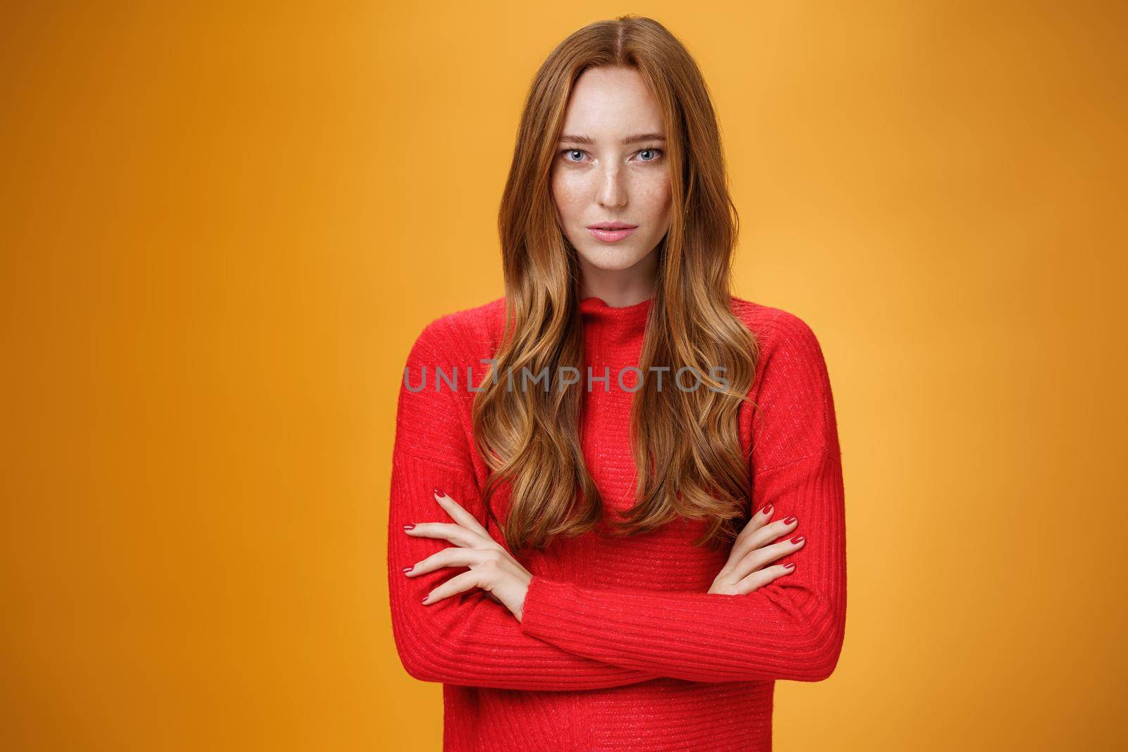 Strong and confident good-looking stylish redhead woman with daring gaze and self-assured attitude holding hands crossed over body looking focused at camera over orange background.