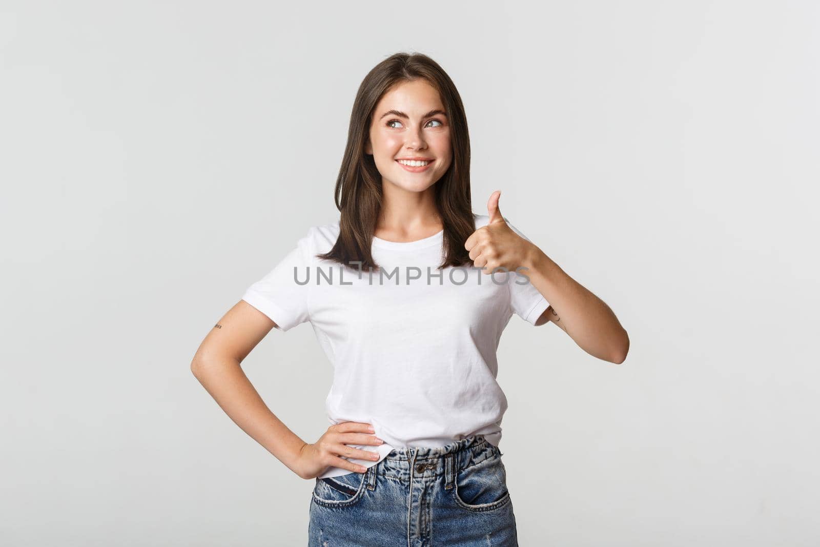 Satisfied smiling young woman showing thumbs-up in approval, looking upper left corner.