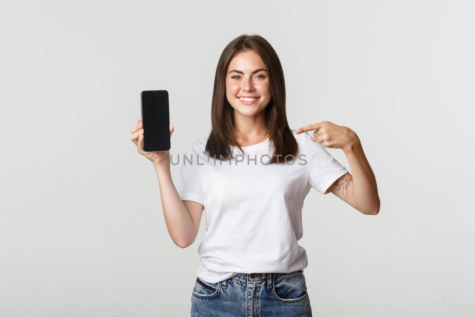 Attractive smiling woman pointing finger at smartphone screen, white background by Benzoix