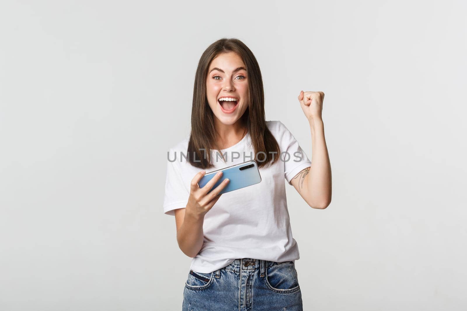 Attractive cheerful brunette girl winning in mobile game, holding smartphone and rejoicing.