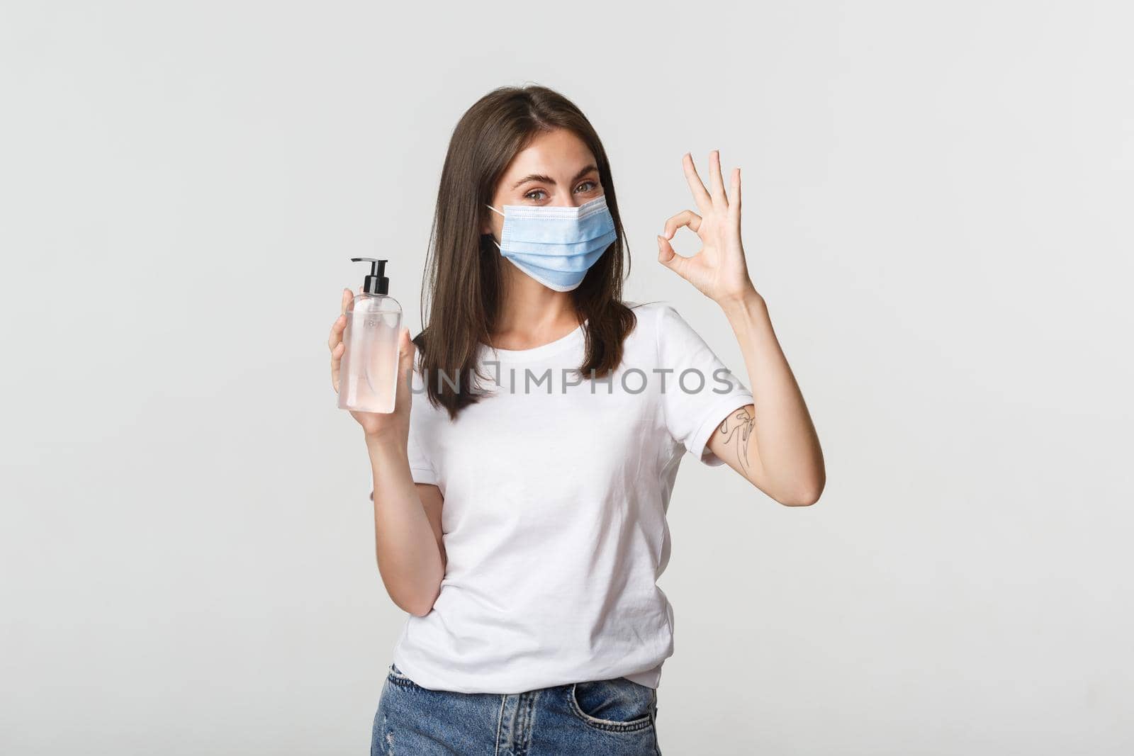 Covid-19, health and social distancing concept. Portrait of smiling brunette girl in medical mask, showing hand sanitizer and okay gesture.