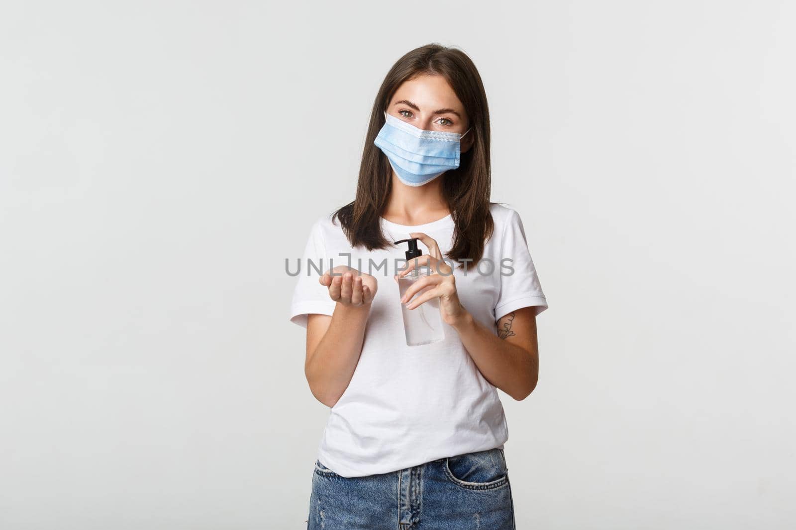 Covid-19, health and social distancing concept. Attractive young brunette woman in medical mask applying hand sanitizer on hand, white background.