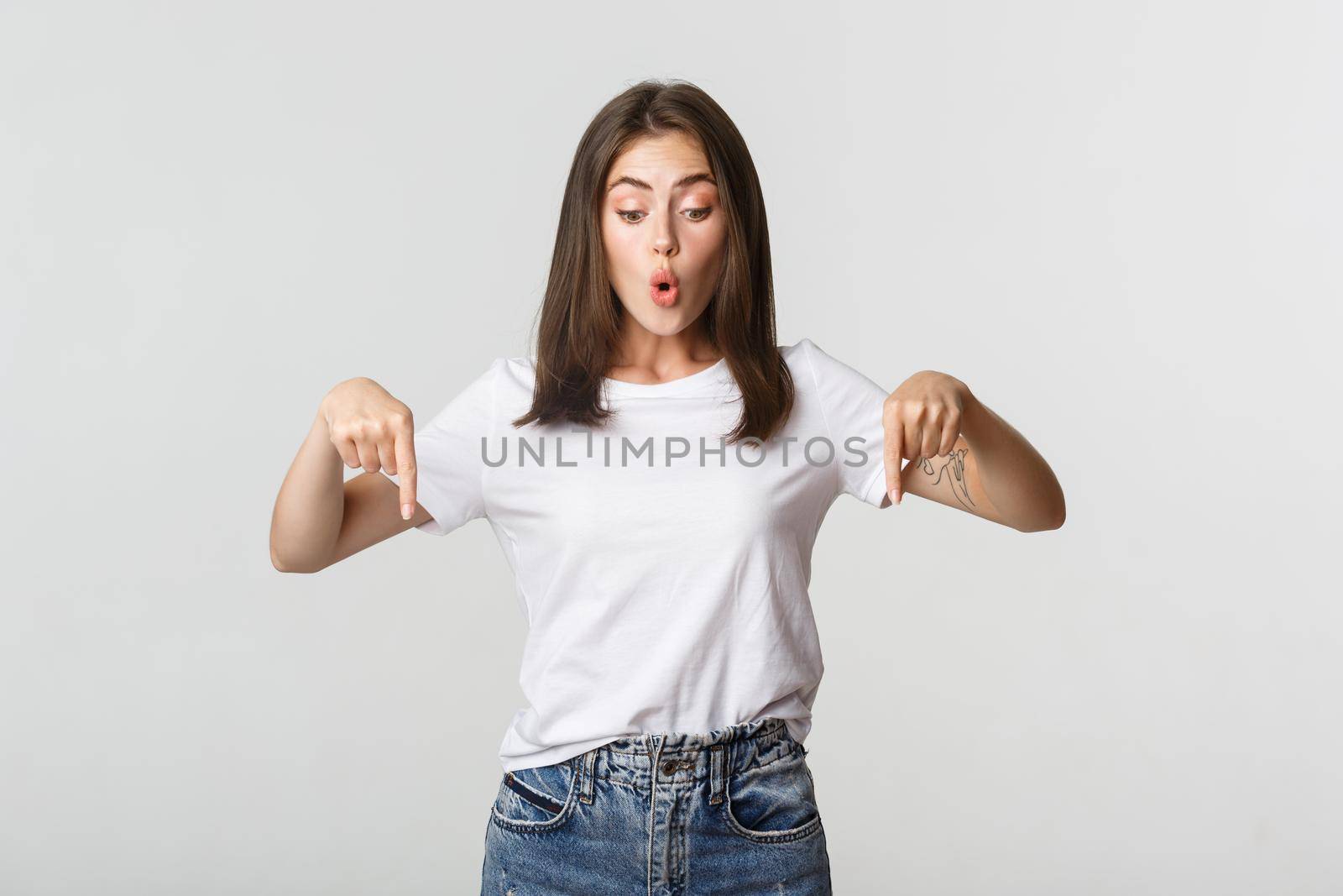 Intrigued attractive girl looking amused and pointing fingers down at logo.